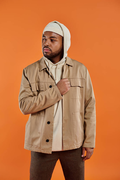 Uomo afro-americano in posa in giacca abbronzata e felpa bianca con cappuccio sullo sfondo vibrante. - Foto, immagini