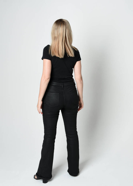Ganzkörperporträt einer schönen blonden Frau in modernem schwarzen Hemd und Lederhose. Selbstbewusst im Stehen vor der Kamera, Silhouette auf weißem Studiohintergrund. - Foto, Bild