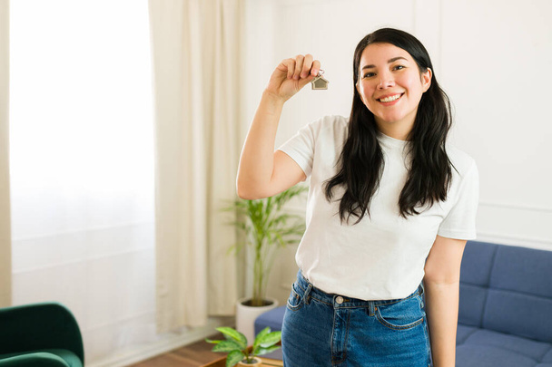 Happy young woman proudly displays new house keys, smiling joyfully in her bright, sunny living room, indicating new homeownership - Fotoğraf, Görsel