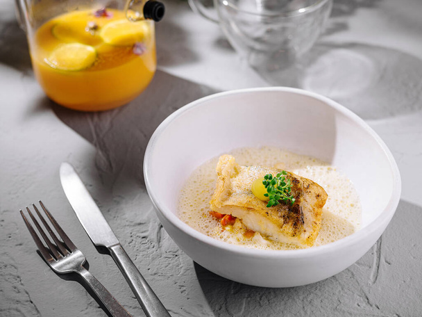 Elegante visfilet geserveerd in een witte kom met een romige saus, vork en mes op een getextureerde tafel - Foto, afbeelding
