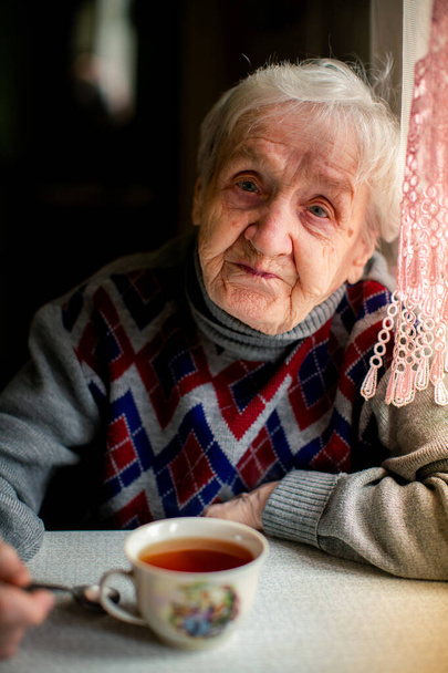 An elderly woman drinks tea by the window, portrait. - Foto, afbeelding