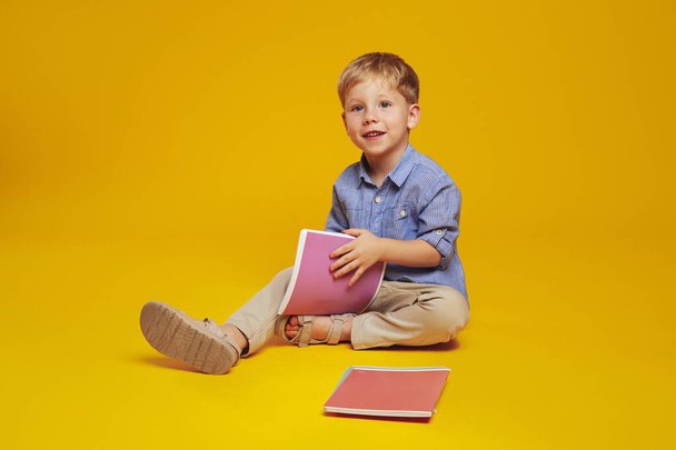 Clever little boy with blonde hair and blue shirt, sitting on studio floor and holding notebooks while looking away, isolated over yellow background. - Foto, Imagen
