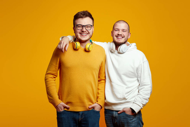 Two cheerful young men with headphones wearing casual clothes, smiling and looking at camera while standing isolated over yellow background. Two cheerful friends embrace each over - Φωτογραφία, εικόνα
