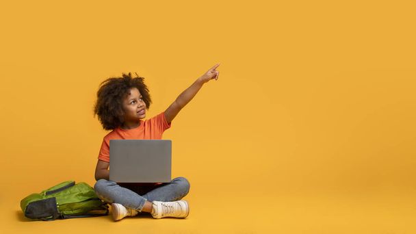 Ein junges afroamerikanisches Mädchen sitzt auf dem Boden, vor sich einen Laptop. Sie zeigt auf etwas auf dem Bildschirm und konzentriert sich intensiv auf die digitalen Inhalte, die angezeigt werden.. - Foto, Bild