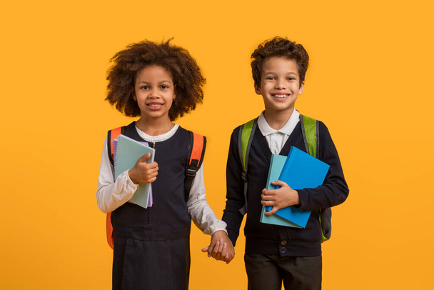 Two African American school children, a boy and a girl, stand hand in hand in front of a bright yellow background. The children are smiling and looking towards the camera - Photo, Image