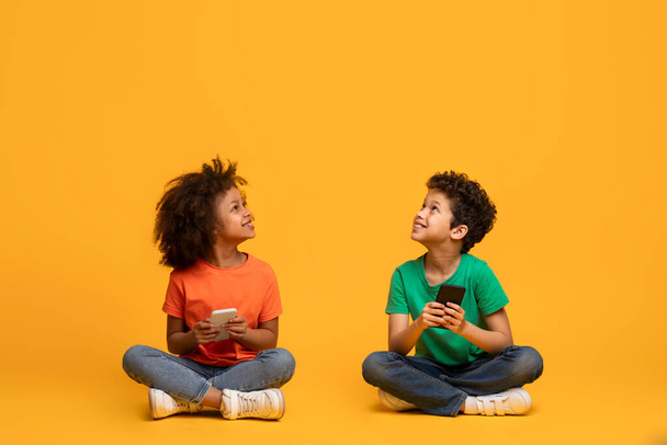 Két afro-amerikai gyerek ül a padlón, belefeledkezve az okostelefonjaikba. Figyelmük kizárólag a képernyőkre összpontosul, ahogy kölcsönhatásba lépnek a körülöttük lévő digitális világgal, utánozzák a teret - Fotó, kép