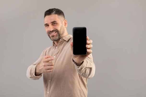 Un hombre barbudo con una camisa beige sonríe mientras sostiene un teléfono inteligente con una pantalla en blanco. El fondo es gris sólido, y el hombre apunta hacia la cámara con su otra mano, maqueta - Foto, Imagen