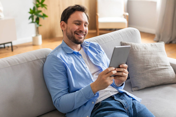 Un uomo è seduto su un divano, assorto nell'uso di un tablet. Si concentra sullo schermo, toccando e scorrendo i contenuti. La stanza è ben illuminata - Foto, immagini