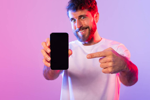 Ein gut gelaunter Mann mit Bart, der ein weißes T-Shirt trägt, zeigt aufgeregt auf einen Smartphone-Bildschirm, während er vor einem leuchtend rosa und lila Neonhintergrund steht.. - Foto, Bild