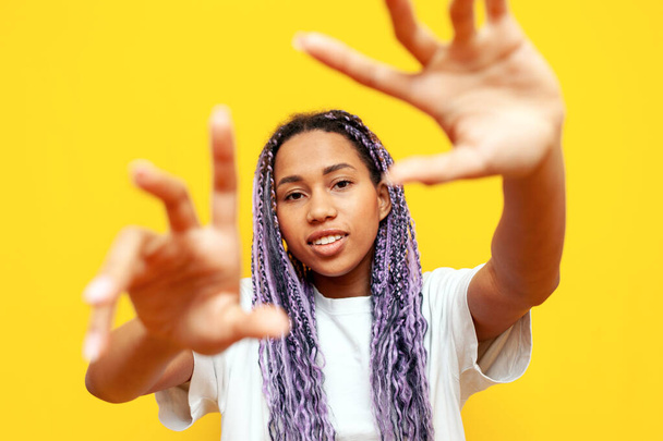 黄色い隔離された背景の上に空の手を示し,保持する着色されたドレッドロックを持つ陽気なアフリカ系アメリカ人女性,紫色のブレードとユニークなヘアスタイルの計画フレームを持つヒップスターガール - 写真・画像