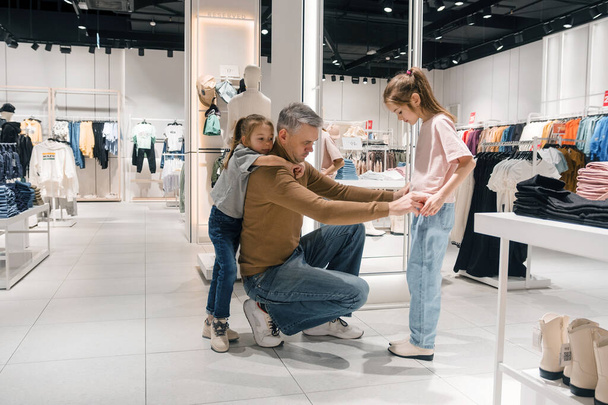Un padre assiste attentamente le sue figlie durante un viaggio di shopping, assicurandosi che trovino i vestiti perfetti in un negozio ben illuminato e spazioso.. - Foto, immagini