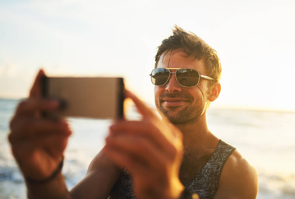 Άνθρωπος, γυαλιά ηλίου και τηλέφωνο στην παραλία για selfie με χαμόγελο, διακοπές στο νησί του Μαυρίκιου. Άνδρας, κινητό και ευτυχία για εικόνα προφίλ στα μέσα κοινωνικής δικτύωσης, ταξίδια ή διακοπές στον ωκεανό κατά το ηλιοβασίλεμα. - Φωτογραφία, εικόνα