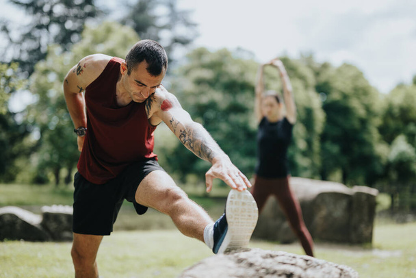 Összpontosított, érett férfi kinyújtja a lábát egy sziklára tetovált karokkal a parkban, miközben egy szőke nő jóga gyakorlatokat végez a háttérben.. - Fotó, kép