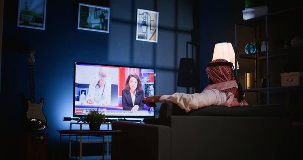 Arabski mężczyzna leżał na kanapie, oglądając wiadomości podczas relaksu w salonie po ciężkim dniu w pracy. Bliski Wschód osoba moafing na kanapie patrząc na kanał telewizyjny w mieszkaniu słabo oświetlone - Zdjęcie, obraz