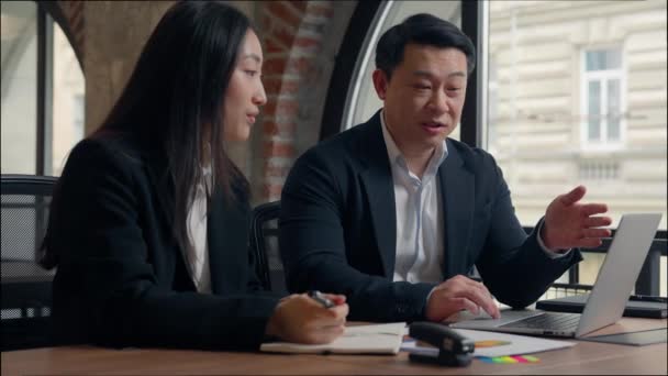 Two Asian business colleagues talking discuss online task computer work Korean man businessman teach using laptop corporate software Japanese Chinese businesswoman intern woman writing notes in office - Filmagem, Vídeo