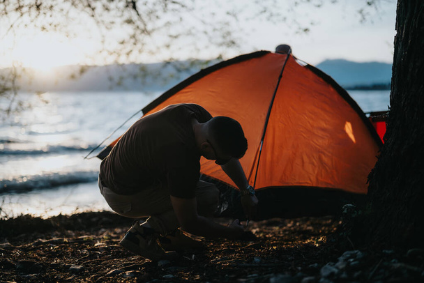 Один человек устанавливает палатку для кемпинга у спокойного озера в сумерках, наслаждаясь мирным открытым сценарием. - Фото, изображение