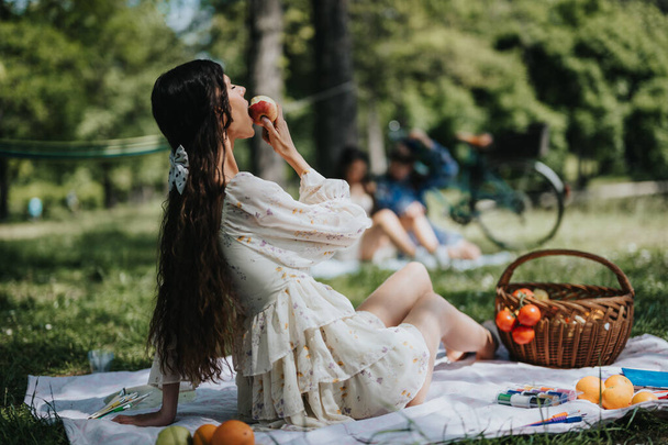 花のドレスを着た若い女性が公園のピクニックブランケットに座り,リラックスして楽しい表情でリンゴを食べます.. - 写真・画像