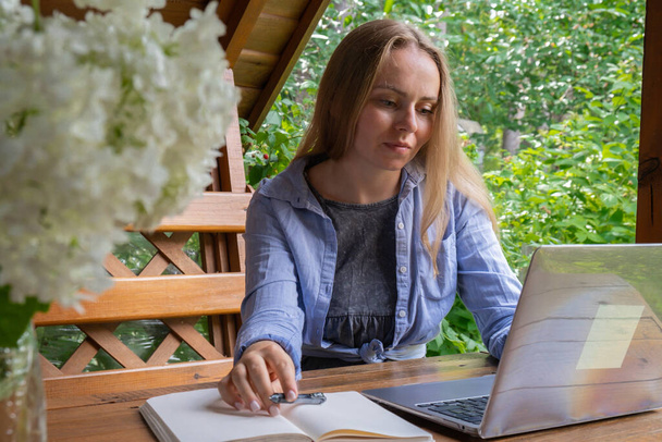 Женщина студентка имеет онлайн урок обучения на открытом воздухе в саду деревянный альков. Блондинка, сидящая снаружи на работе на ноутбуке и получающая видеозвонок. Единство с природой - Фото, изображение