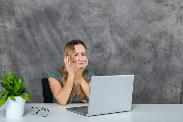 A young woman with lovely blond hair and an elegant gray dress creates various poses in front of a laptop, with a vibrant green flower adding a touch of nature to the scene. - Foto, Bild