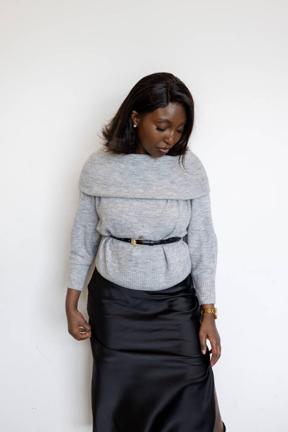 This image features a Black woman posing against a white background, dressed in a stylish gray sweater and a black skirt. She is looking downwards with a thoughtful expression, adding a contemplative - Foto, imagen