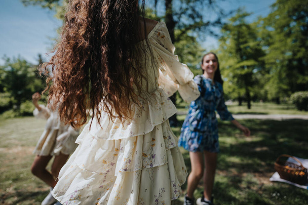 Mujeres jóvenes vestidas con vestidos florales vistos desde atrás, deleitándose con alegría despreocupada mientras juegan en un parque soleado, encarnando la esencia de la primavera. - Foto, Imagen