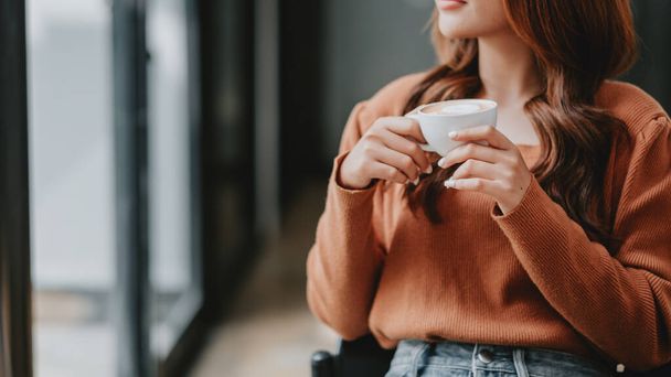 A woman is sitting in a chair with a cup of coffee in her hand. She is wearing an orange sweater and has long brown hair. Concept of relaxation and comfort - Foto, imagen