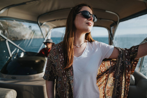 Une femme joyeuse avec des lunettes de soleil étend les bras sur un bateau, sentant la liberté pendant qu'un ami navigue, reflétant une journée de vacances parfaite sur le lac. - Photo, image