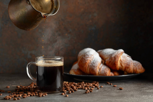Coffee is poured into a mug. Croissants and black coffee on a kitchen table. Focus on a foreground. - Photo, Image