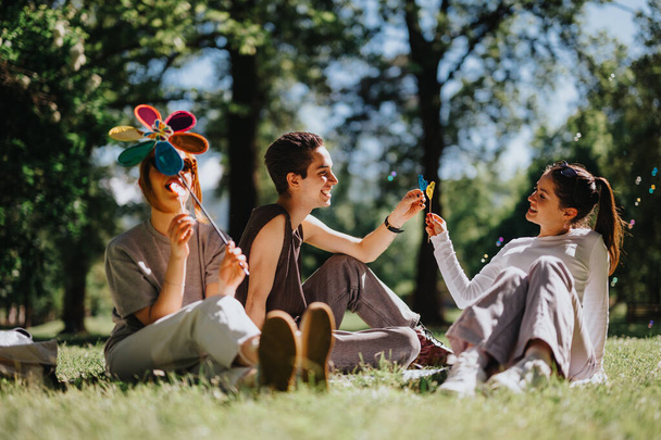 Three young friends sit on the grass in a sunny park, sharing a joyful moment while playing with vibrant, colorful lollipops. - Foto, immagini