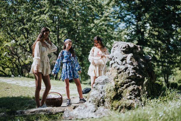 Three young females in stylish summer dresses gathering by a rock fountain in a lush, sunny park. - Photo, Image