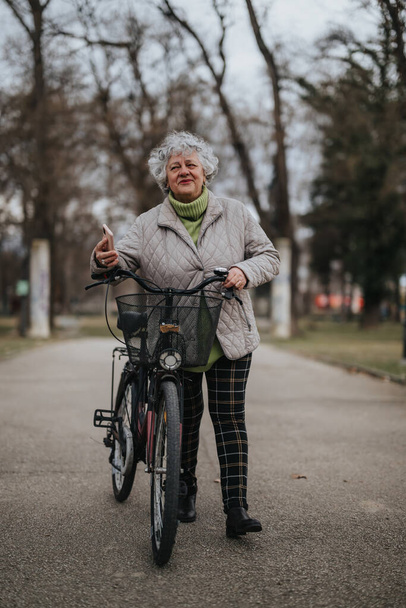 Dojrzała kobieta z rowerem stojąca w parku, ciesząca się przyrodą i aktywnym stylem życia w sezonie jesiennym. - Zdjęcie, obraz