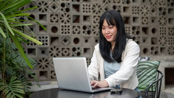 カジュアルな服を着た若いポジティブなアジア人女性がカフェで遠隔操作を行い,ラップトップコンピュータで働き,ラップトップのキーボードをタイプし,屋外のテーブルに座っている.. - 写真・画像