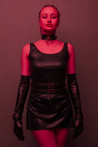 Beautiful young woman in a leather dress and bondage set posing on red light backgound - Φωτογραφία, εικόνα