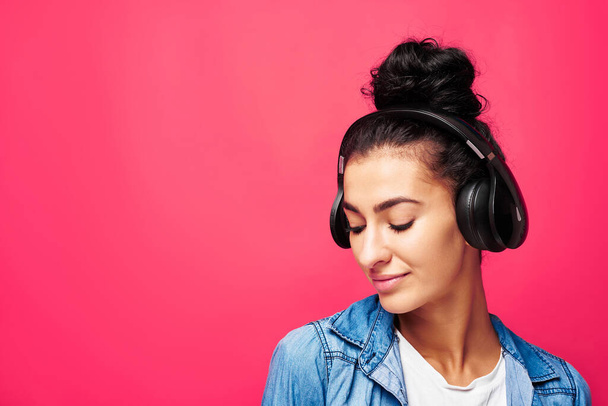 Stüdyoda duran ve kulaklık takan çekici bir Arap kadının portresi. Müzik dinlemekten zevk alıyorum. Pembe arka planda izole edilmiş günlük elbiseli bir kız. - Fotoğraf, Görsel