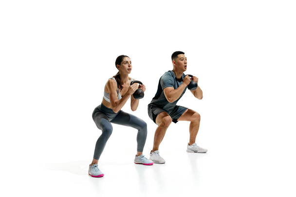 Man en vrouw oefenen met kettlebells in squat positie tegen witte studio achtergrond. Atletische bewegingen. Concept van mensen in de sport, gezonde levensstijl, teamwork, motivatie. Steun - Foto, afbeelding