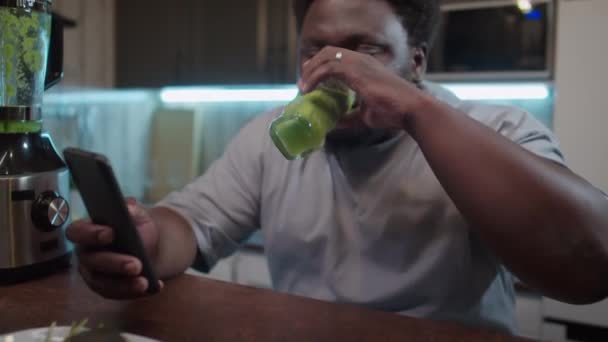 キッチンテーブルに座っている肥満の若い黒人男性,自宅で夕食中に携帯電話で緑色の滑らかさとテキストメッセージを飲んで - 映像、動画