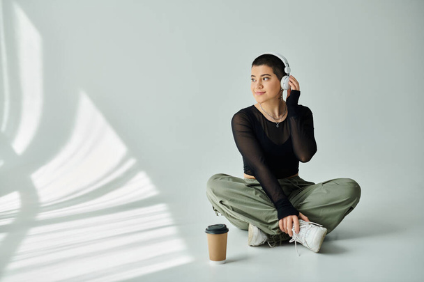 Une jeune femme aux cheveux courts s'assoit sur le sol, dégustant une tasse de café en tenue décontractée élégante sur un fond gris. - Photo, image
