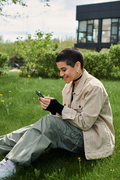 Стильна жінка з коротким волоссям, що сидить на траві, зосереджена на екрані смартфона. - Фото, зображення