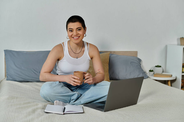 Una joven con el pelo corto está sentada en su cama, enfocada en la pantalla de su computadora portátil. - Foto, imagen