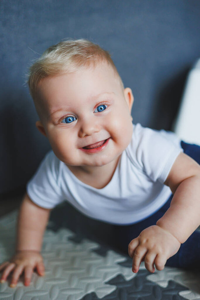 A blue-eyed baby boy in a white bodysuit and blue pants learns to sit. Self-development of the child. The baby is sitting on the floor and smiling - Photo, Image