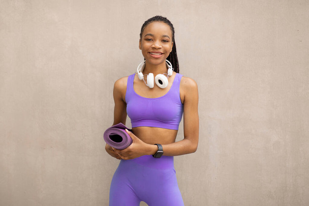 Urban sports. Happy african american female in sports uniform resting after workout outdoors, holding fitness mat, standing over beige wall - 写真・画像