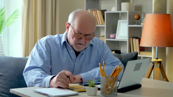 Un hombre de negocios anciano, canoso con una camisa azul, en su escritorio, revisando datos de negocios, trabajando en una computadora y una calculadora. Está muy concentrado. Movimiento lento. - Imágenes, Vídeo