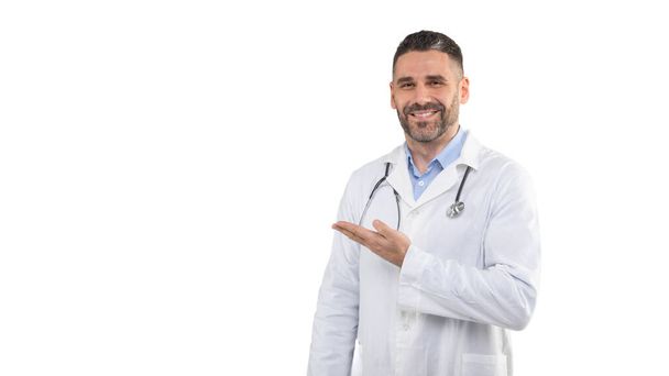 Mies lääkäri, yllään valkoinen labratakki ja stetoskooppi, hymyilee kun hän elehtii sivulle. Tausta on tavallinen, hyvin valaistu studio, korostaen hänen ammatillinen pukea, kopioi tilaa - Valokuva, kuva