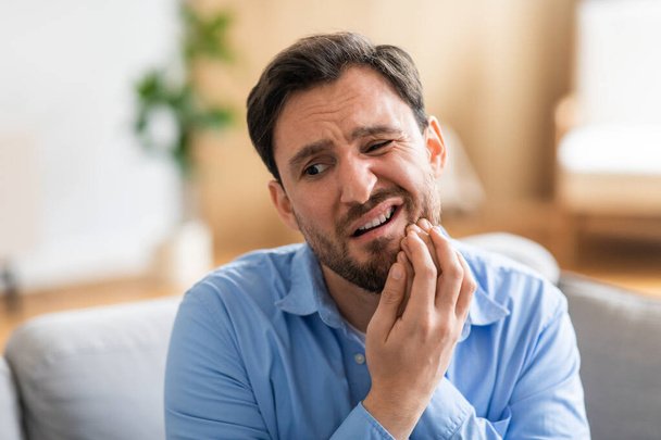 Un uomo a disagio è seduto su un divano, tenendo il viso dolorante a causa di un mal di denti. La sua espressione rivela la sua sofferenza mentre cerca di alleviare il dolore. - Foto, immagini