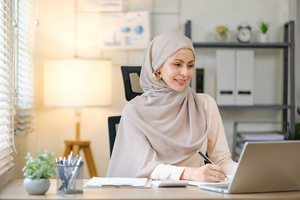 Egy fejkendőt viselő nő ül egy asztalnál egy laptoppal és egy noteszral. Mosolyog és élvezi a munkáját. - Fotó, kép