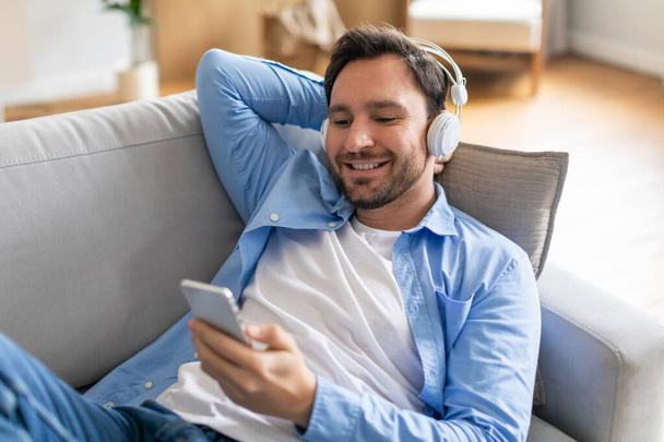 Muž sedí na gauči a poslouchá hudbu ve svém telefonu. Vypadá uvolněně a zapojený do hudby přehrávání přes sluchátka. - Fotografie, Obrázek