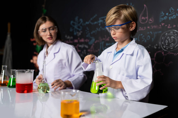 Вчитель підтримує школяра в лабораторії, вони носять лабораторне пальто і окуляри, стоять і експериментують з хімії в класі STEM. Студентський язик твердий до жовтої рідини в склянці. Руйнування. - Фото, зображення