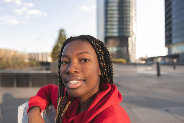 Fröhliche junge Frau in rotem Kapuzenpulli, die draußen lächelt, während im Hintergrund städtische Gebäude stehen. - Foto, Bild