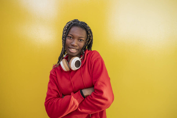 Zelfverzekerde jonge vrouw in een rode hoodie met koptelefoon om haar nek, glimlachend met armen gekruist tegen een gele achtergrond. - Foto, afbeelding