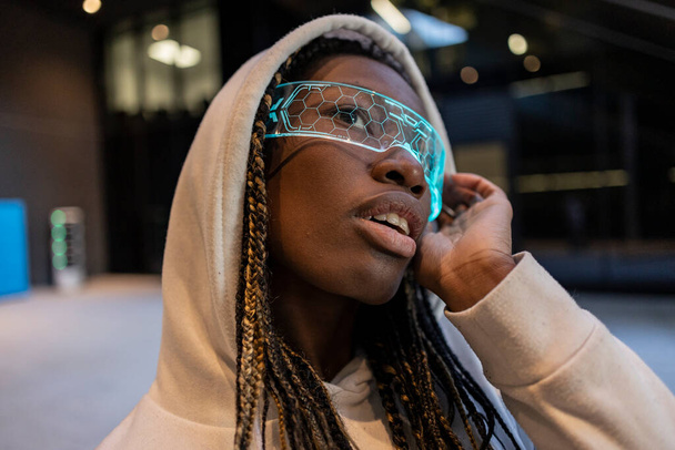 Nahaufnahme einer jungen Frau mit neonfuturistischer Brille und Kapuzenpullover, die in einer urbanen Umgebung nachdenklich wirkt. - Foto, Bild